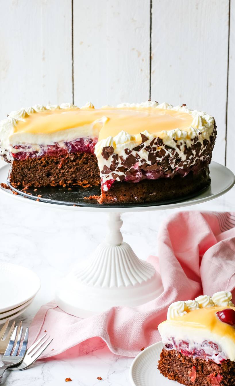 Eierlikörtorte mit Kirschen angeschnitten auf einer Tortenplatte, davor ein Stück des Torte auf einem Teller