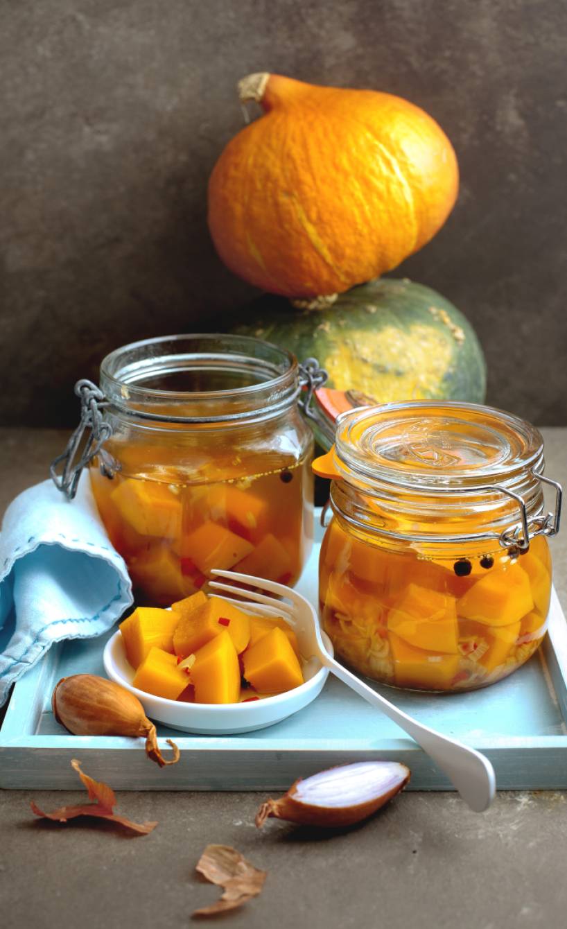 Eingelegter Kürbis in zwei Weckgläsern auf einem Tisch umgeben von Zutaten