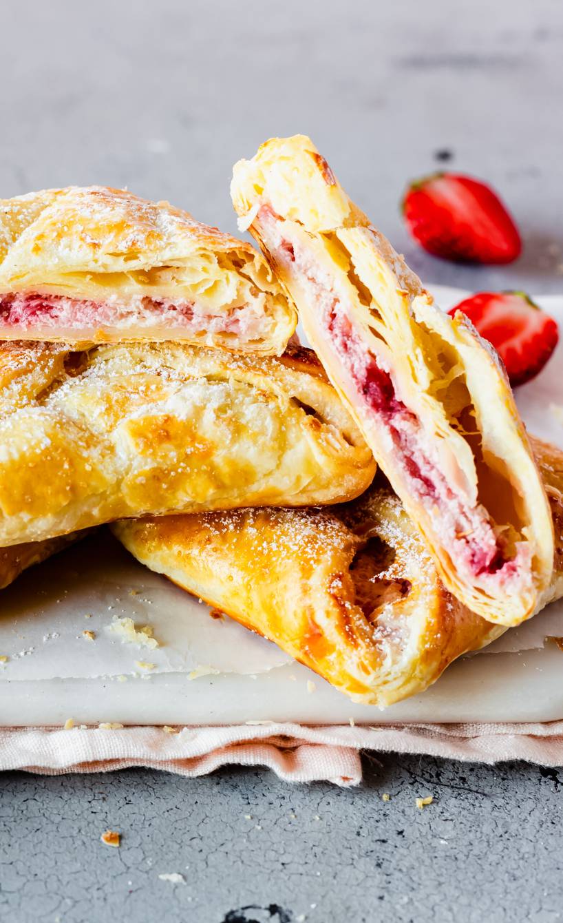 Fünf Blätterteigtaschen gefüllt mit Erdbeeren und Ricotta auf einem Teller, eine halbiert mit gut sichtbarer Füllung 