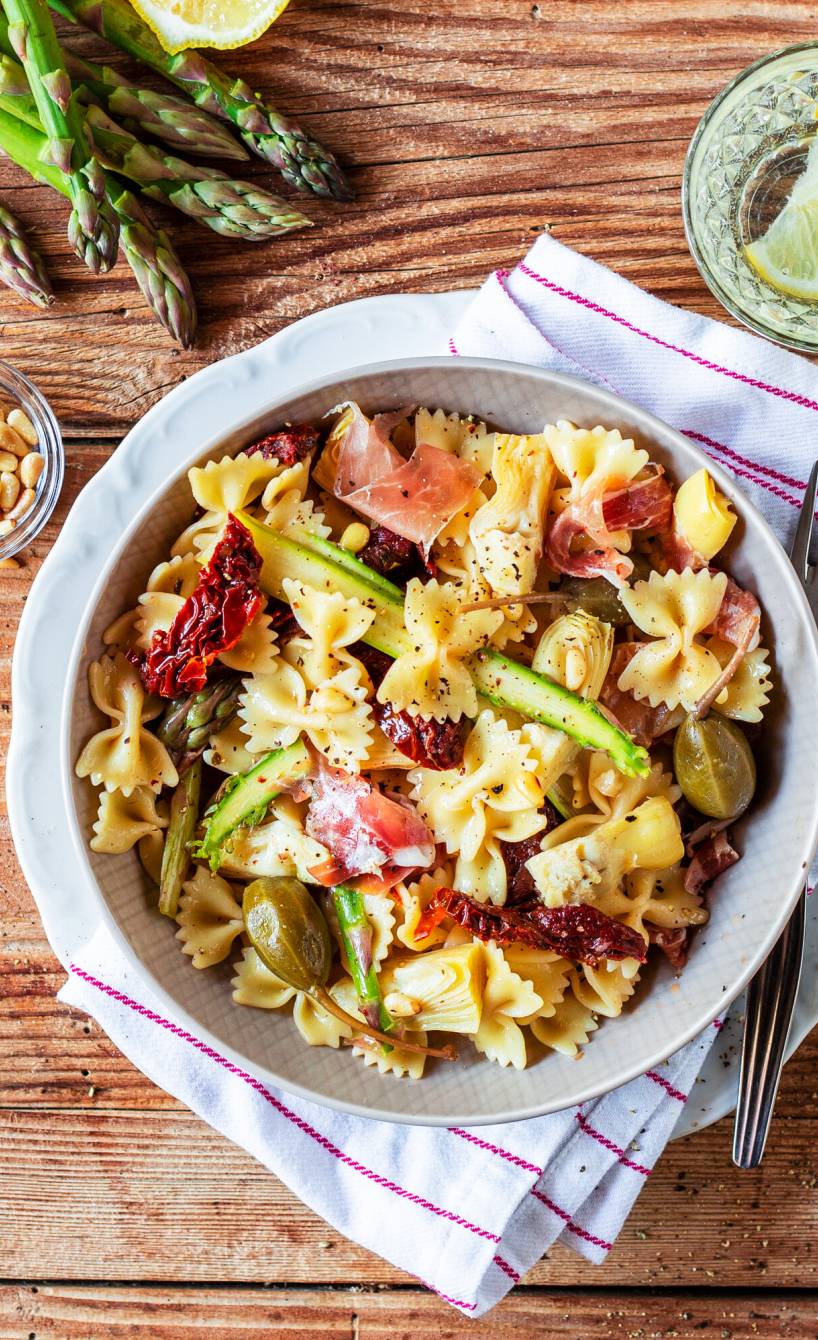 Nudelsalat mit Farfalle, grünem Spargel, Artischocken und Schinken angerichtet in einer Salatschüssel auf einem Holztisch