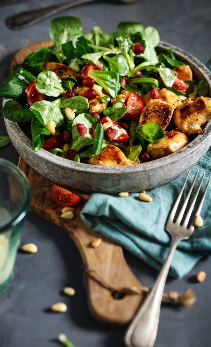 Feldsalat mit gebratenen Hähnchenstücken getoppt von Pinien- und Granatapfelkernen, daneben Parmesandressing