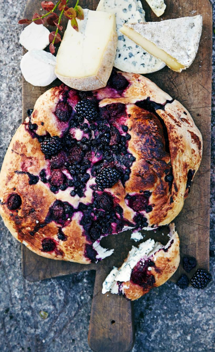 Focaccia mit Brombeeren, Blaubeeren und Rosmarin auf einem Holzbrett, daneben verschiedene Käsestücke