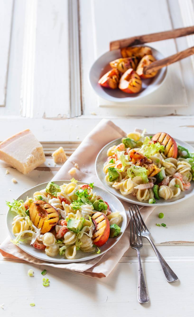 Pastasalat mit Flusskrebsfleich auf einem Teller getoppt von gegrillten Nektarinenstücken und Avocado