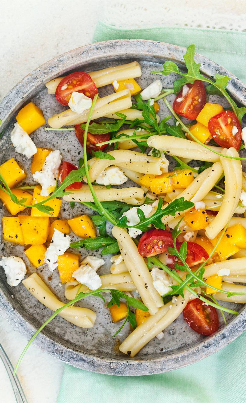 Pastasalat auf einem Teller mit Mangostücken, Schafskäse und Tomaten, daneben Besteck auf einem Tisch