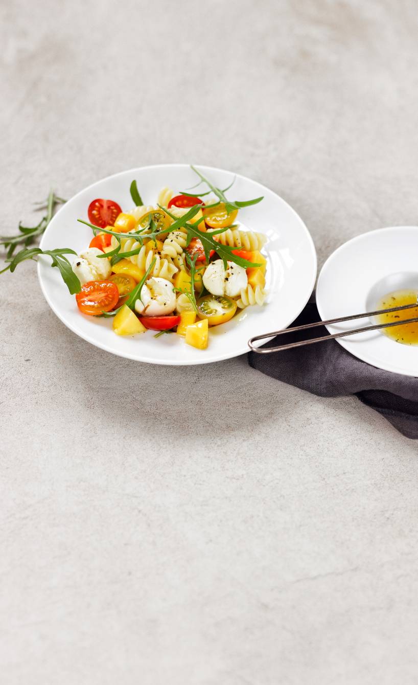 Pastasalat mit Tomaten und Mangostücken auf einem Teller, daneben Dressing in einer Schüssel