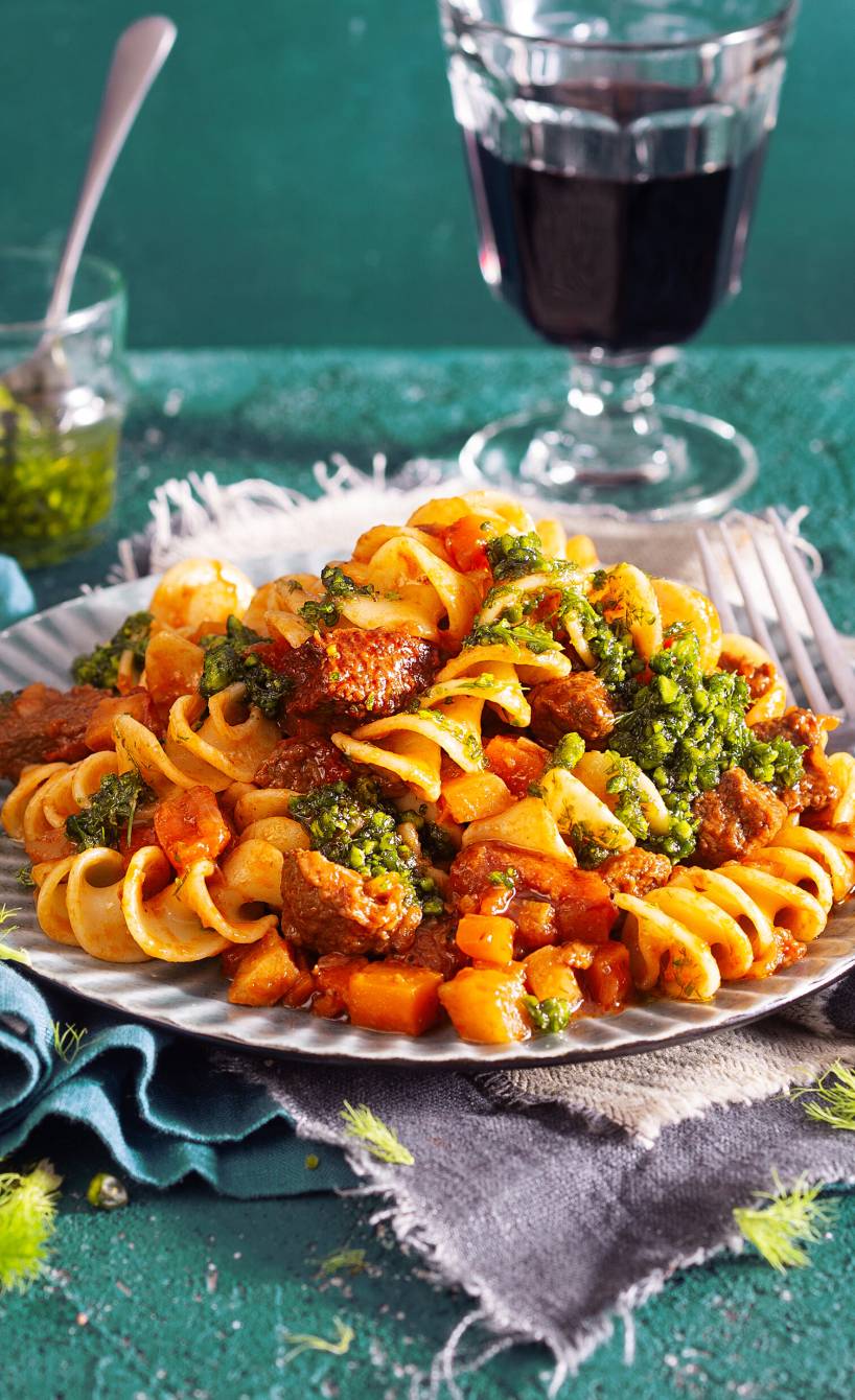 Kalbsragout mit Fusilli auf einem Teller getoppt von Fenchel-Pistazien-Pesto, daneben ein Glas Rotwein auf einem Tisch