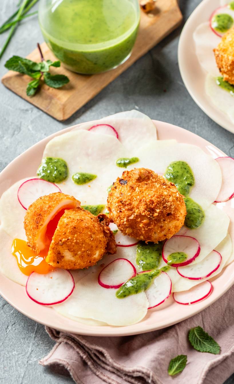 Gebackene Eier in Nusspanade angerichtet auf zwei Tellern auf Kohlrabi-Radieschen-Carpacchio, daneben Dressing in einer Karaffe