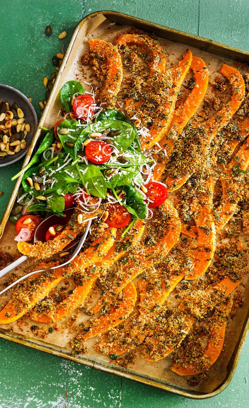 Gebackene Kürbisspalten mit Parmesankruste auf einem Backblech, daneben eine Schale Nüsse