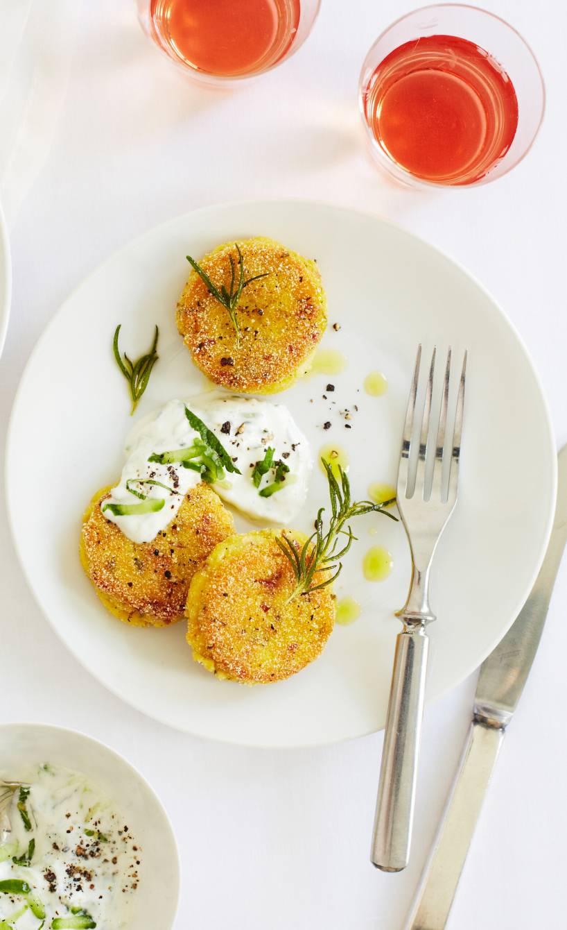 Mehrere gebratene Polenta-Bratlinge mit Schinken und Rosmarin angerichtet auf einem Teller mit Gurken-Minz-Dip