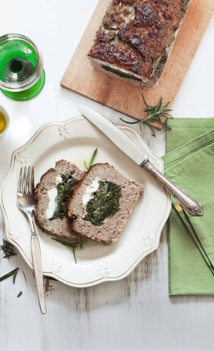 Zwei Stücke Hackbraten gefüllt mit Spinat und Ziegenkäse auf einem Teller, der kastenförmige Rest auf einem Holzbrett