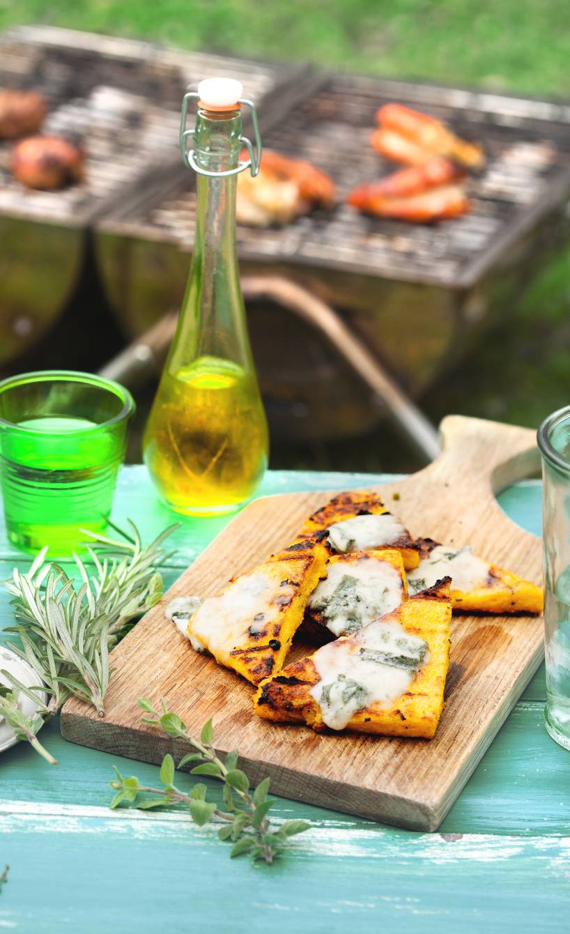 Gegrillte Polentaecken auf einem Holzbrett getoppt mit Gorgonzola, daneben eine Getränkekaraffe auf einem Tisch