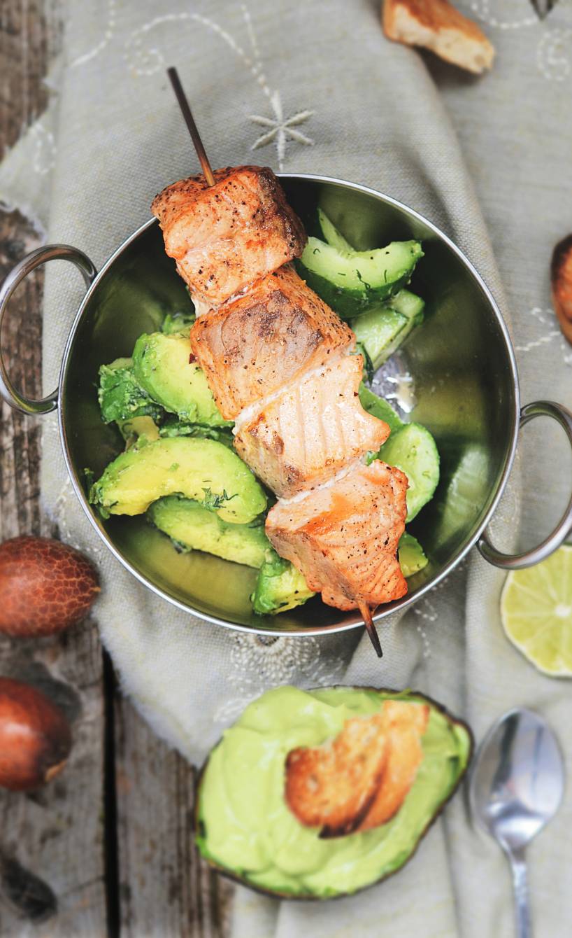 Gegrillter Lachs auf einem Spieß auf Gurkensalat, daneben Avocadodip und halbierte Avocados