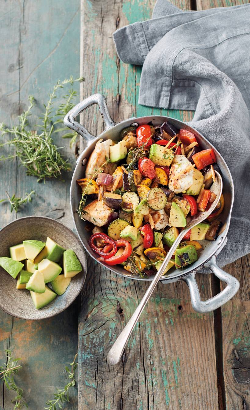Pfanne mit verschiedenen Gemüsesorten und Hähnchenstücken auf einem Tisch, daneben Avocadostücke und frische Kräuter in Schälchen