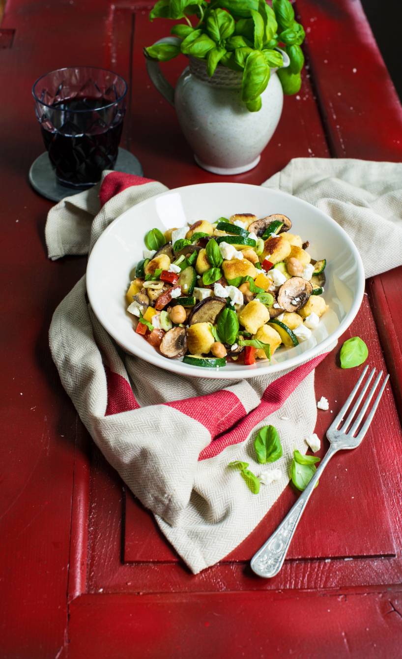 Gnocchi-Gemüse-Pfanne in einem tiefen Teller getoppt mit Feta auf einem Tisch, daneben frische Kräuter