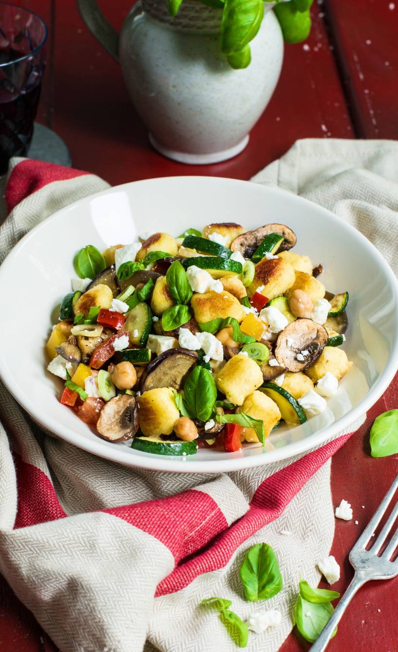 Gnocchi-Gemüse-Pfanne in einem tiefen Teller getoppt mit Feta auf einem Tisch, daneben frische Kräuter