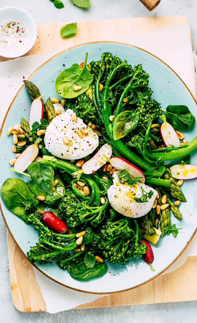 Gemüsesalat mit Spinat, Spargel und Brokkolini getoppt von Mozzarella, Dressing und Pinienkernen auf einem Teller