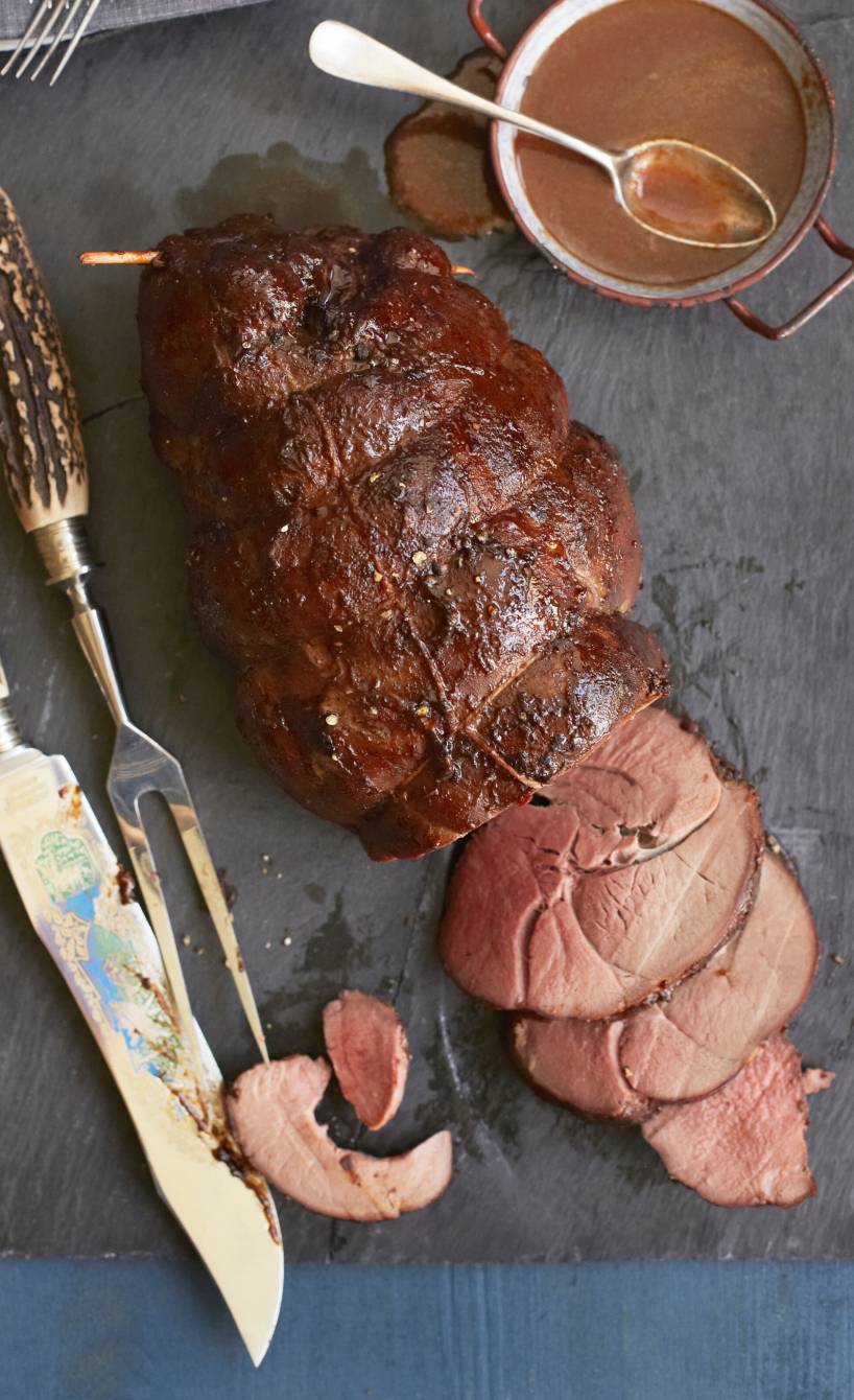 Geschmorte Rehkeule angeschnitten auf einer Servierplatte, daneben dunkle Soße in einer Sauciere