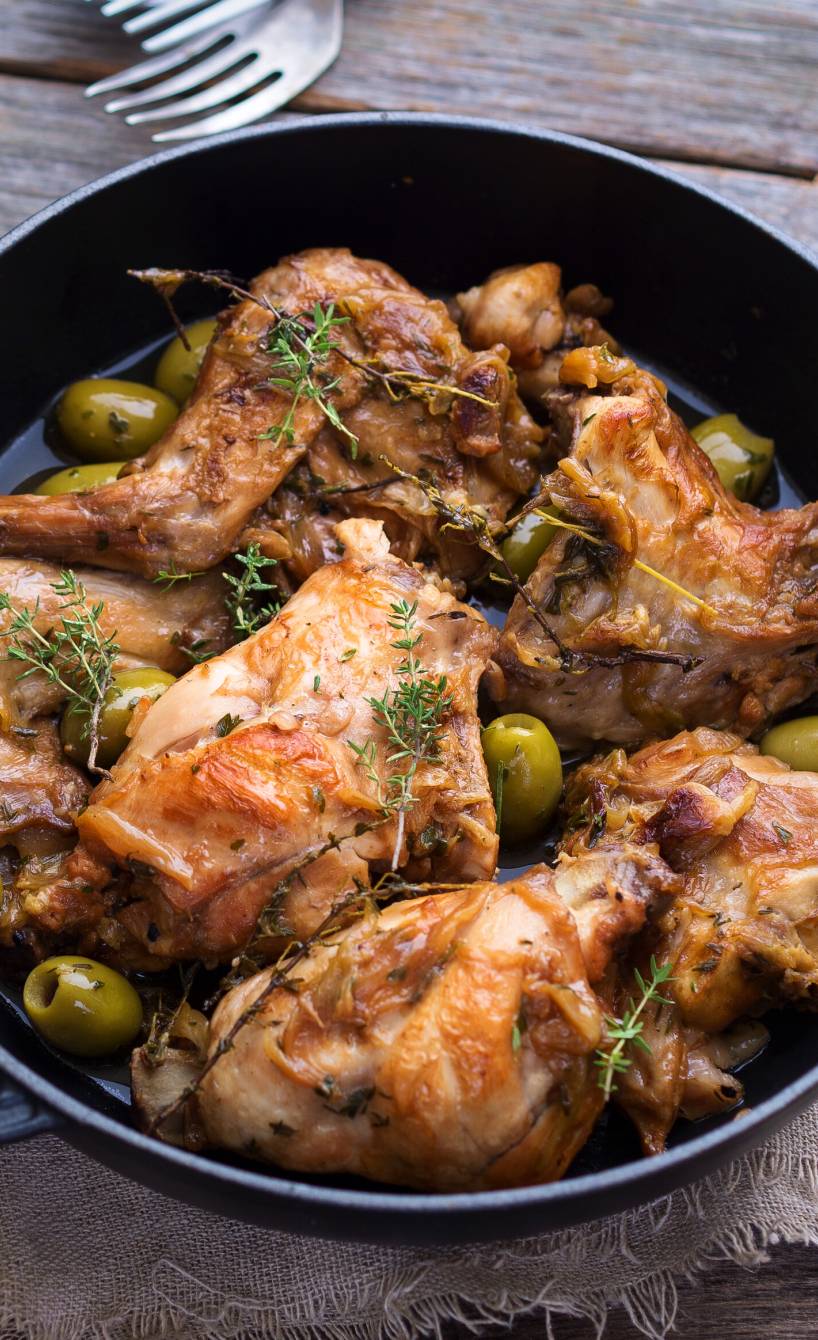 Kaninchenkeulen in einem Schmortopf mit Oliven und Gewürzen getoppt, daneben ein paar Stücke Ciabatta