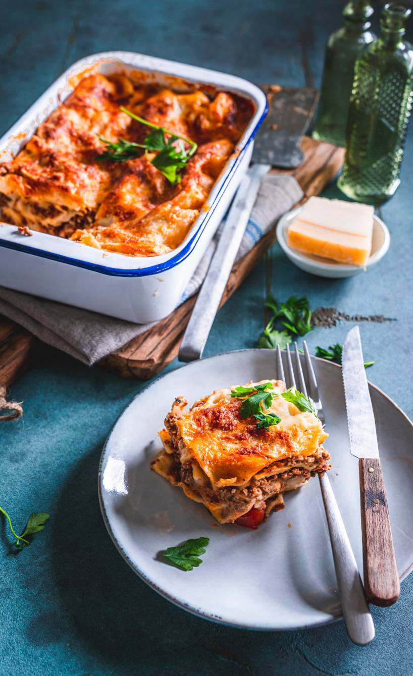 Glutenfreie Lasagne in einer Auflaufform, daneben ein Stück auf einem Teller und Parmesan