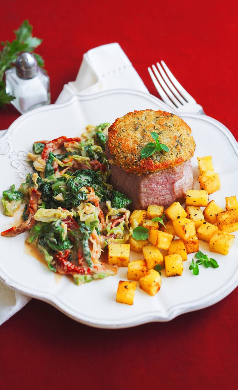 Gratiniertes Kalbsmedaillon mit Kräuterhaube auf einem Teller mit Kartoffelwürfeln und Wirsinggemüse in Sauce