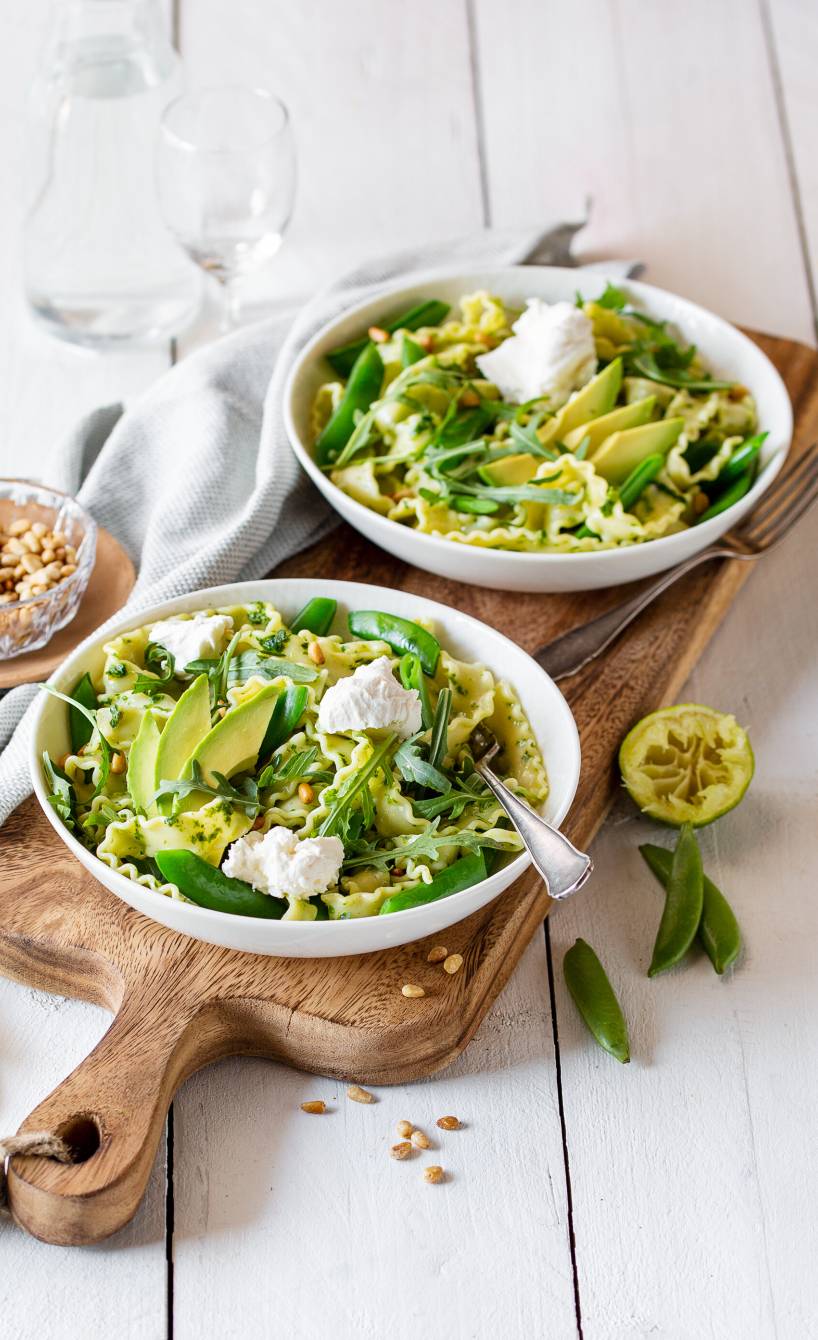 Mafaldine-Salat mit Basilikumpesto-Dressing auf zwei Tellern getoppt von Avcoadostücken, Mascarponeklecksen und Pinienkernen