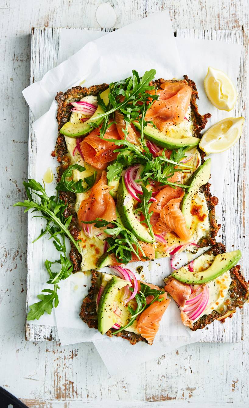 Rechteckige Low-Carb-Pizza mit Grünkohl-Käseboden und Räucherlachs-Avocado-Belag angeschnitten auf einem Holzbrett