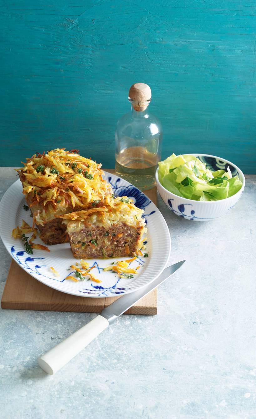 Hackbraten mit Kartoffelhaube angeschnitten auf einer Platte, daneben eine Schale mit grünem Salat und Olivenöl