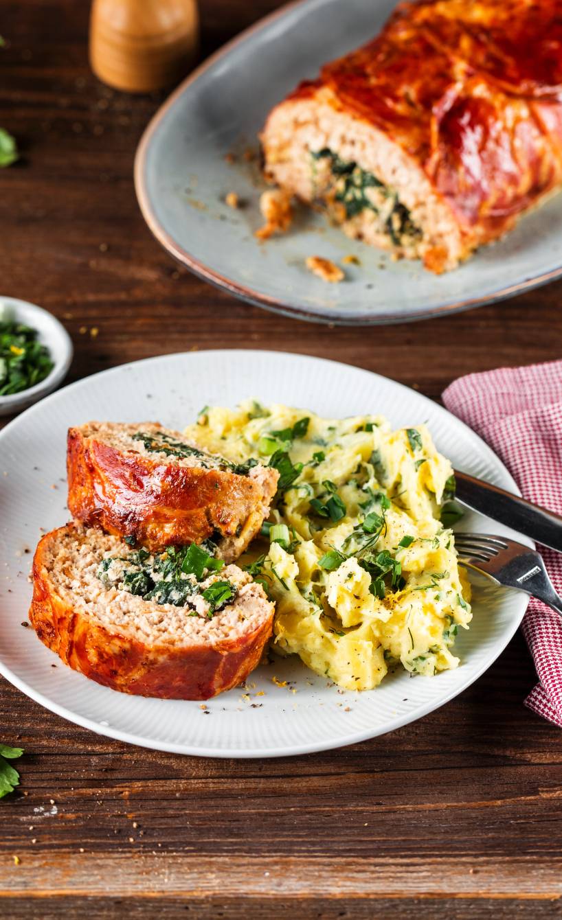 Ein Stück Hackbraten mit Mangold-Ricotta-Füllung, dahinter der restliche Hackbraten im Ganzen auf einem Holztisch
