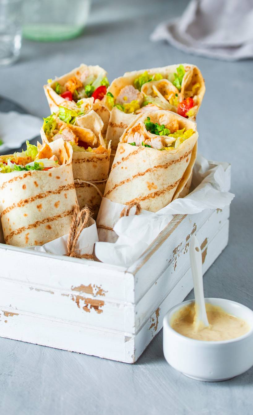 Hähnchen-Wraps nebeneinander in einer kleinen Holzkiste, daneben ein Schälchen mit Caesar-Salad-Creme