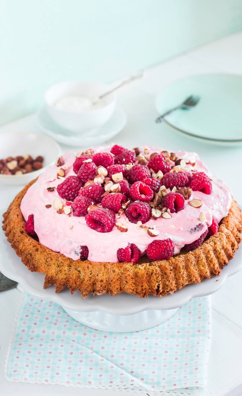 Selbstgebackener Haselnusskuchen getoppt von mit Himbeer-Mascarponecreme, Himbeeren und Haselnusskernen auf einer Kuchenplatte