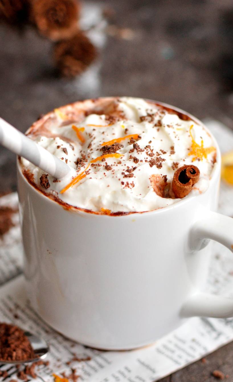 Heiße Schokolade mit Sahne in einer Tasse auf einem Tisch, daneben Zimtstangen und Orangen