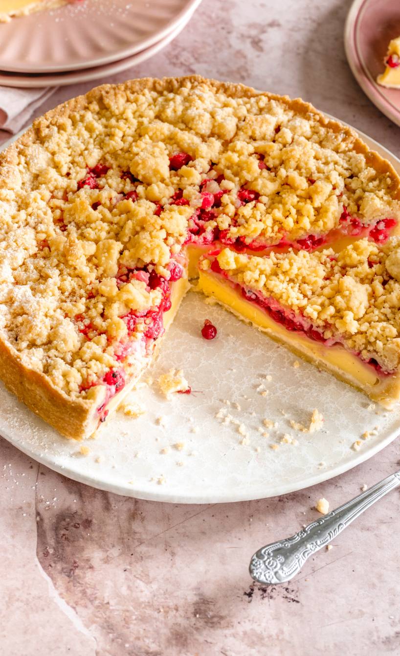 Johannisbeerkuchen mit Pudding und Streuseln angeschnitten auf einer Kuchenplatte, daneben zwei Stücke auf Tellern