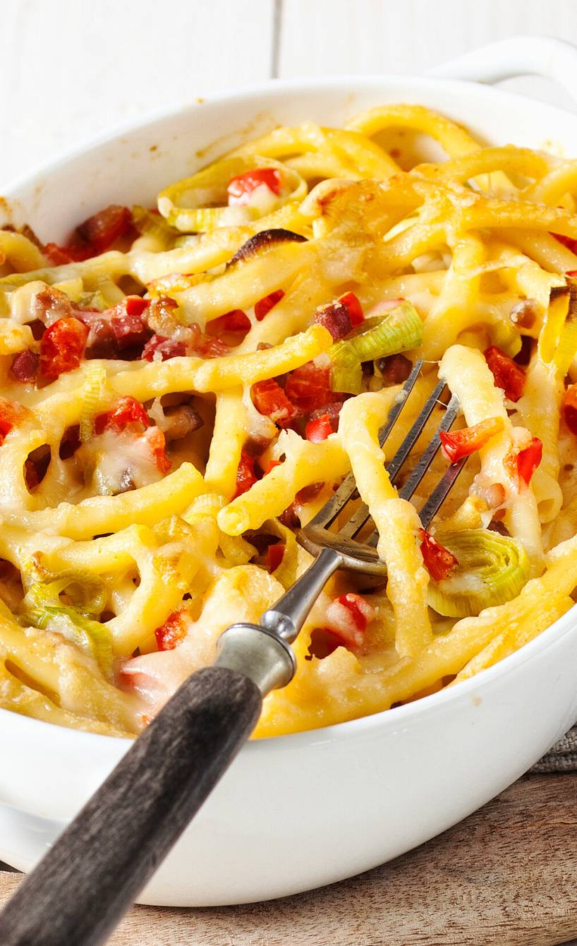 Käse-Makkaroni mit Speck, Porree und Paprika in einer Auflaufform auf einem Holzbrett, daneben eine Käsereibe