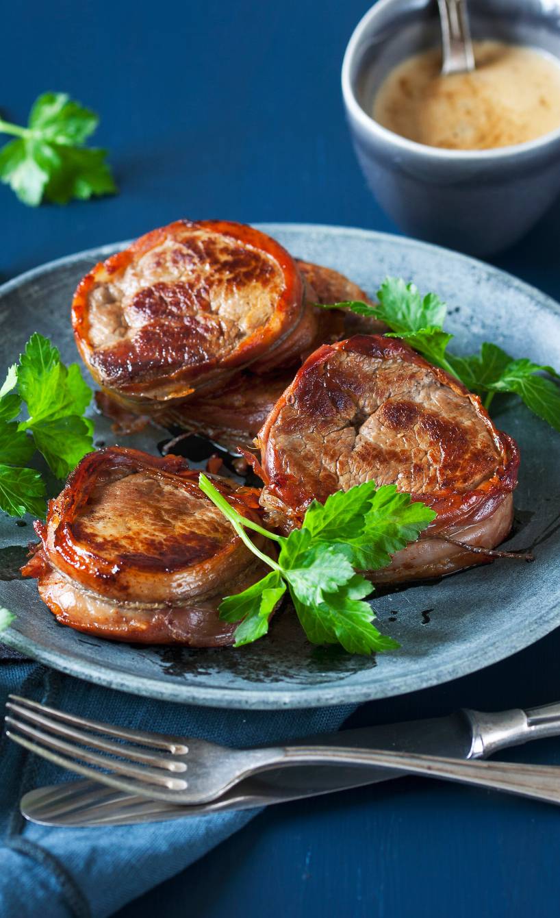 Drei Kalbsmedaillons umhüllt mit Parmaschinken auf einem Teller, daneben Sahnesoße in einer Schale