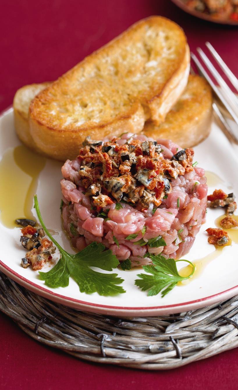 Kalbstartar mit Tomaten-Oliven-Topping auf einem Vorspeisenteller angerichtet, daneben eine Scheibe geröstetes Ciabatta