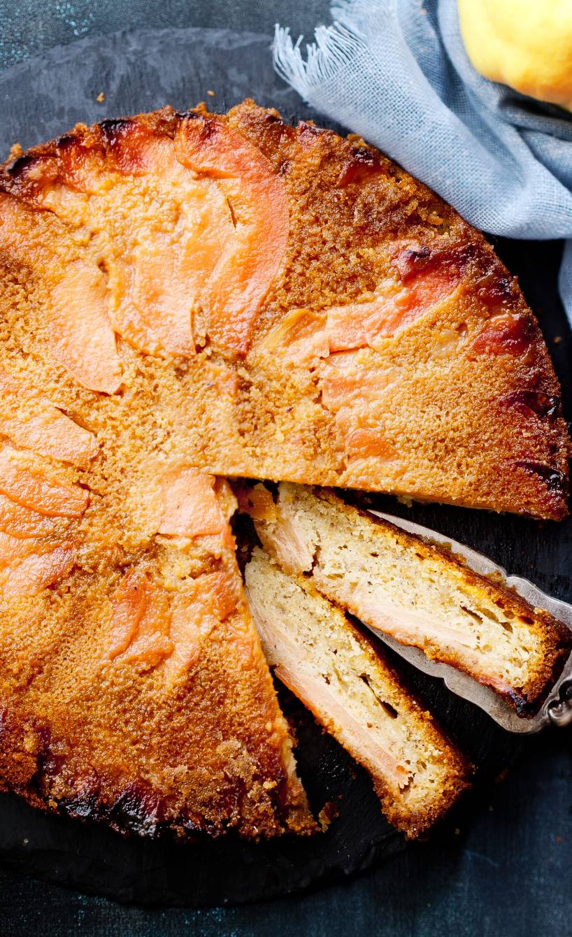 Runder Quittenkuchen angeschnitten auf einer Kuchenplatte, daneben zwei Quitten auf einem Tisch