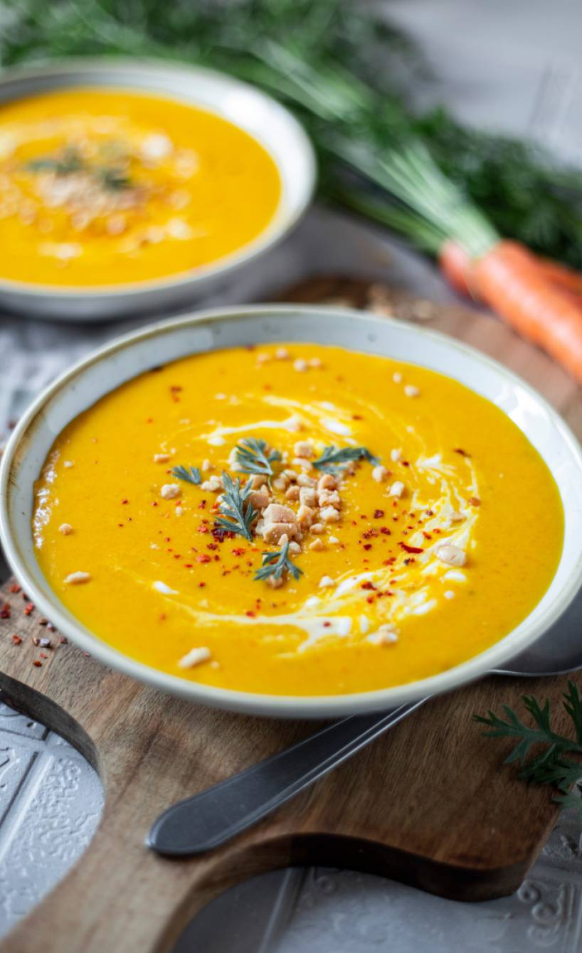 Karotten-Cremesuppe in zwei tiefen Tellern auf einem Tisch, daneben Bundmöhren