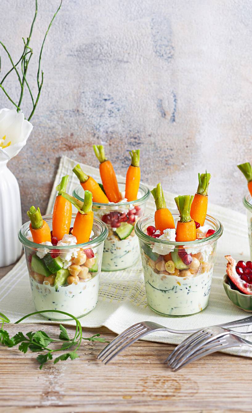 Schichtsalat mit Karotten, Kichererbsen, Feta, Joghurtdressing und Granatapfelkernen in vier Gläsern auf einem festlich dekorierten Tisch