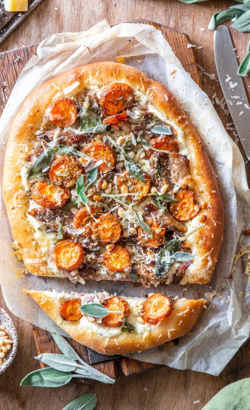 Ovale Pizza mit Entenbrust, Karotten und Pilzen belegt angeschnitten und mit Pinienkernen daneben