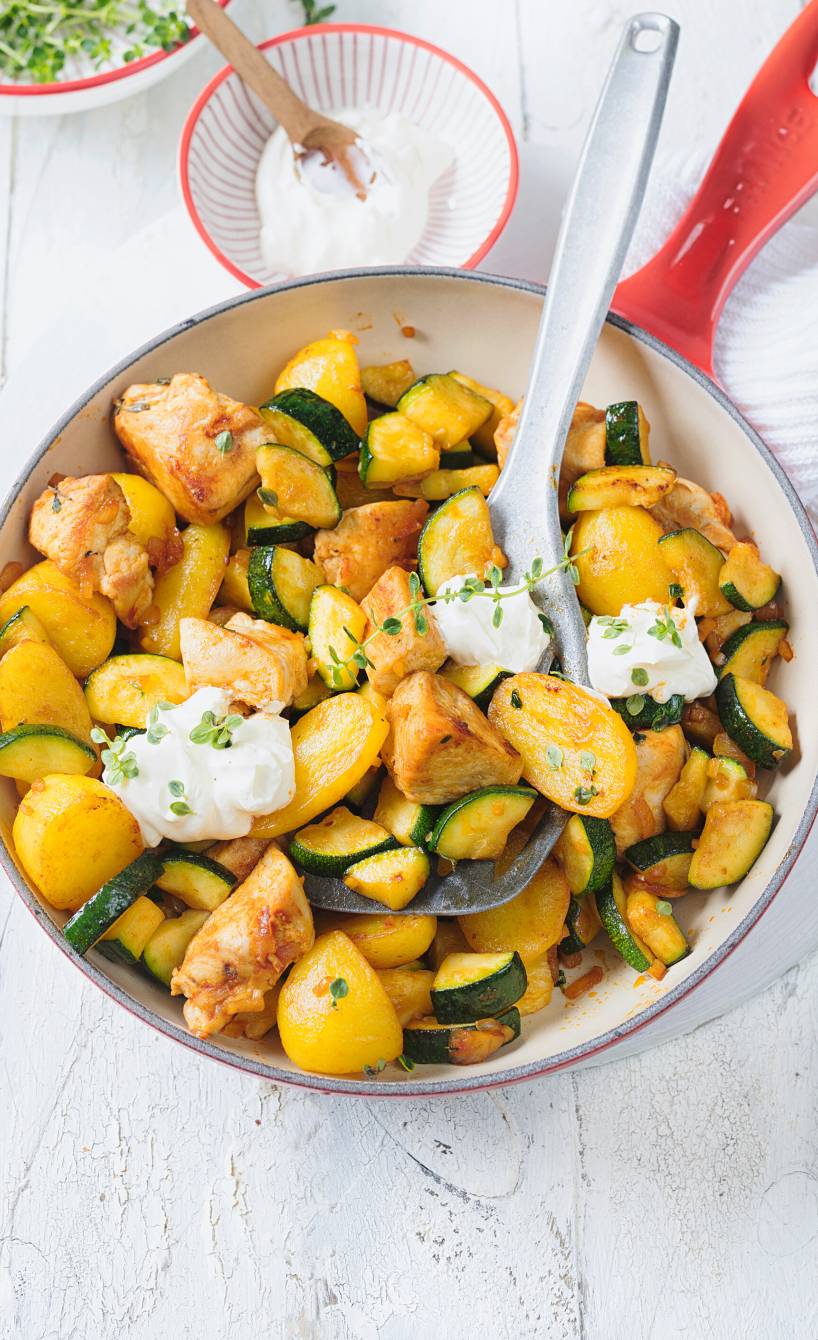 Pfanne mit Hähnchen, Kartoffeln und Zucchini getoppt von Schmand-Klecksen und frischen Kräutern auf einem Tisch
