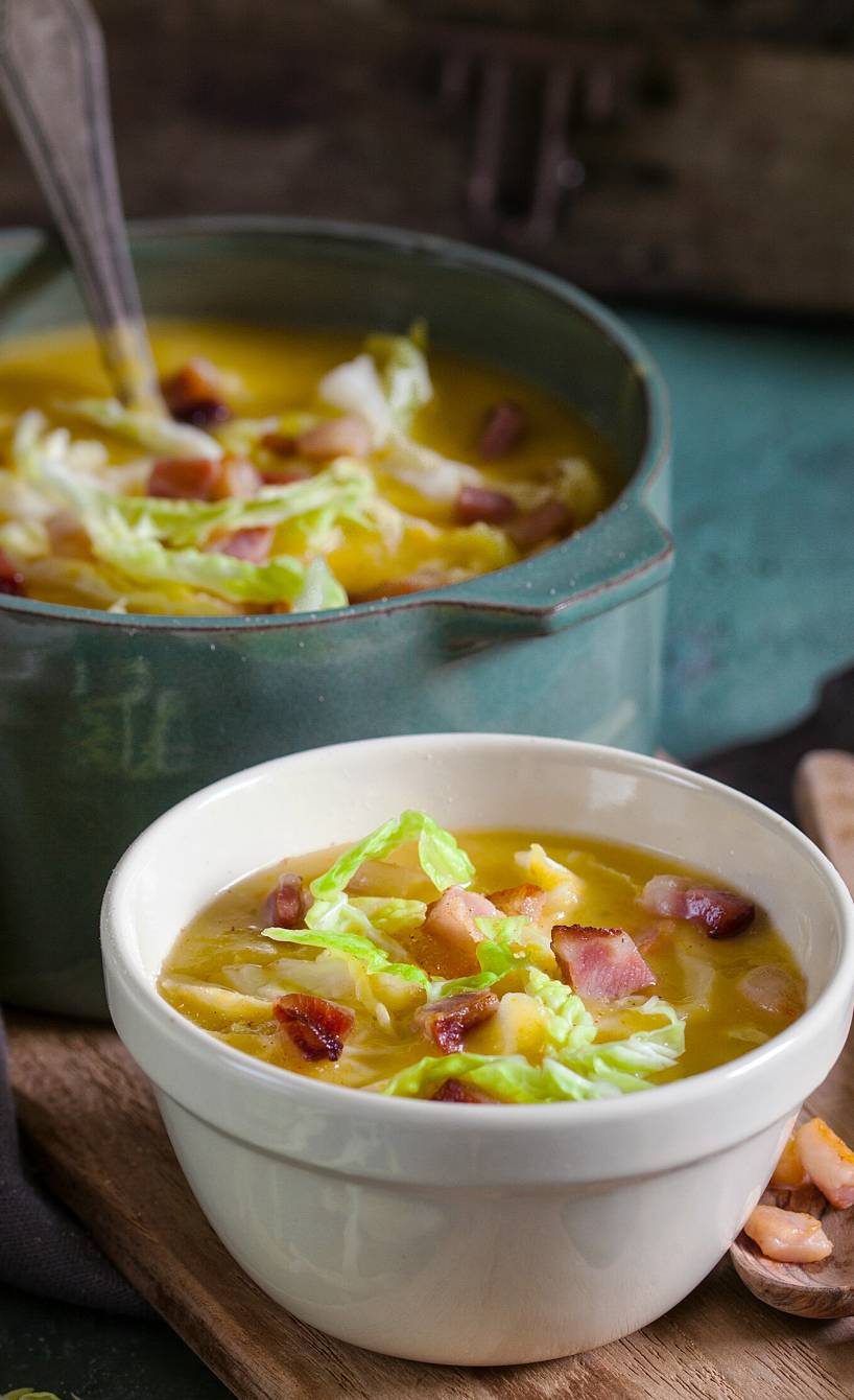 Kartoffelsuppe mit Wirsing getoppt mit Speckwürfeln in einer Suppenschüssel, davor eine Suppenschale