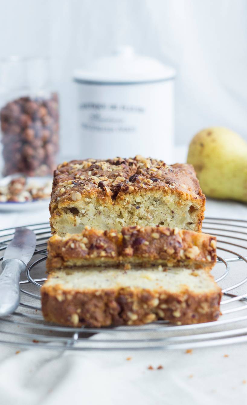 Angeschnittener Kartoffelkuchen aus der Kastenform auf einem Kuchengitter, daneben Haselnüsse und Birnen auf einem Tisch