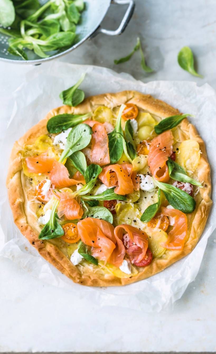 Runde Kartoffelpizza belegt mit Räucherlachs, Tomaten und Feldsalat auf einem Stück Backpapier