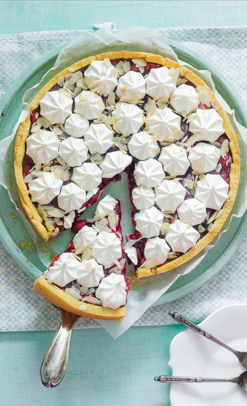 Runder Kirschkuchen mit Amaretto und Baisertüpfelchen angeschnitten auf einer Kuchenplatte auf einem Holztisch von oben