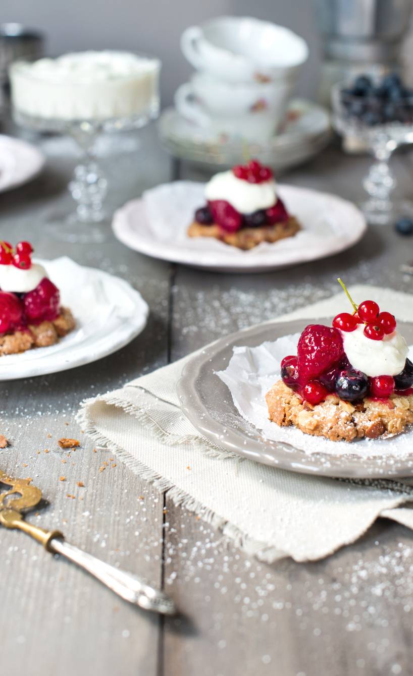 Mehrere Beeren-Törtchen mit Haferflocken-Mandelboden und Crème fraîche-Topping auf Kuchentellern auf einem Tisch