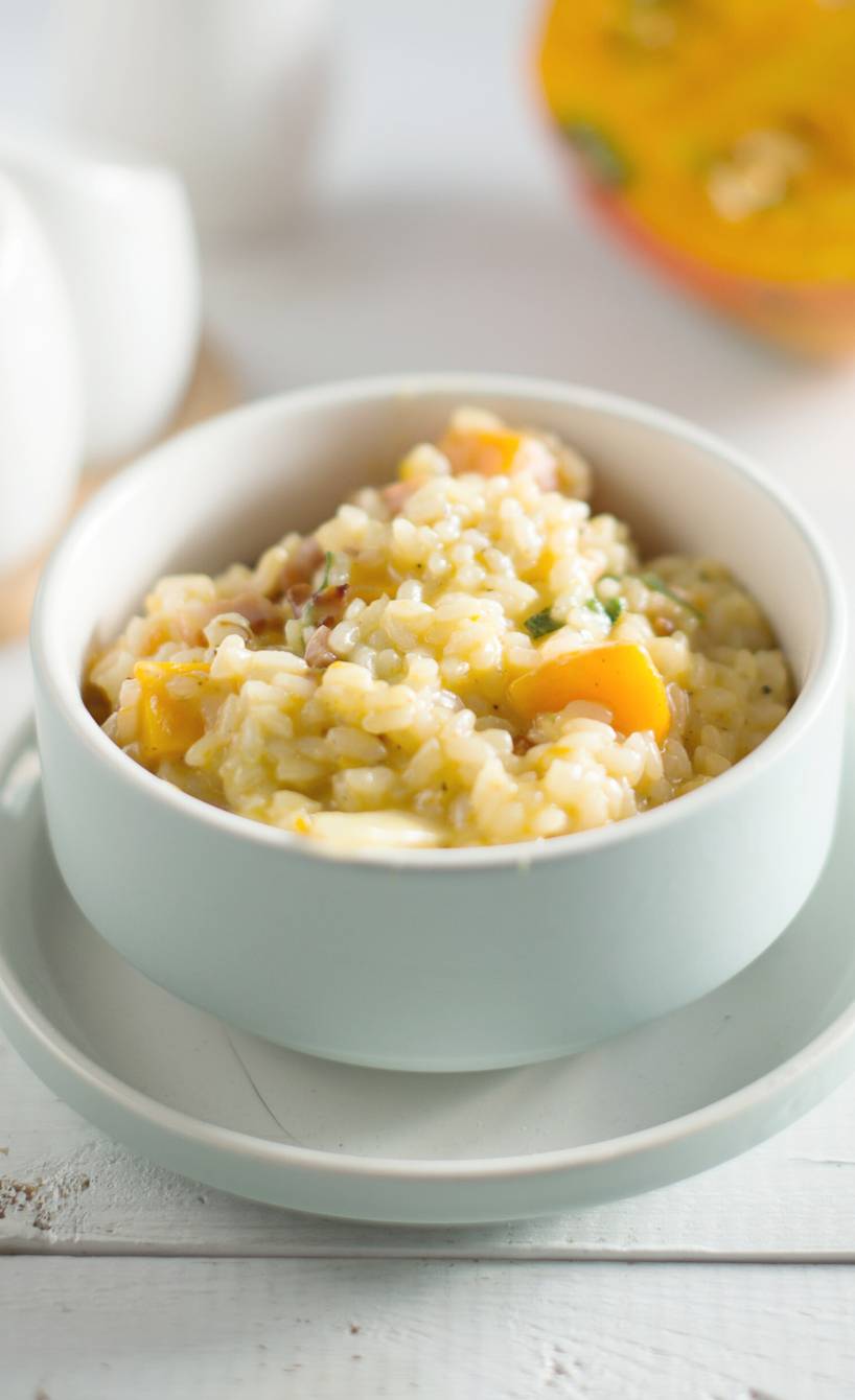 Kürbisrisotto in einer Schale auf einem Tisch, dahinter ein Stück Hokkaidokürbis