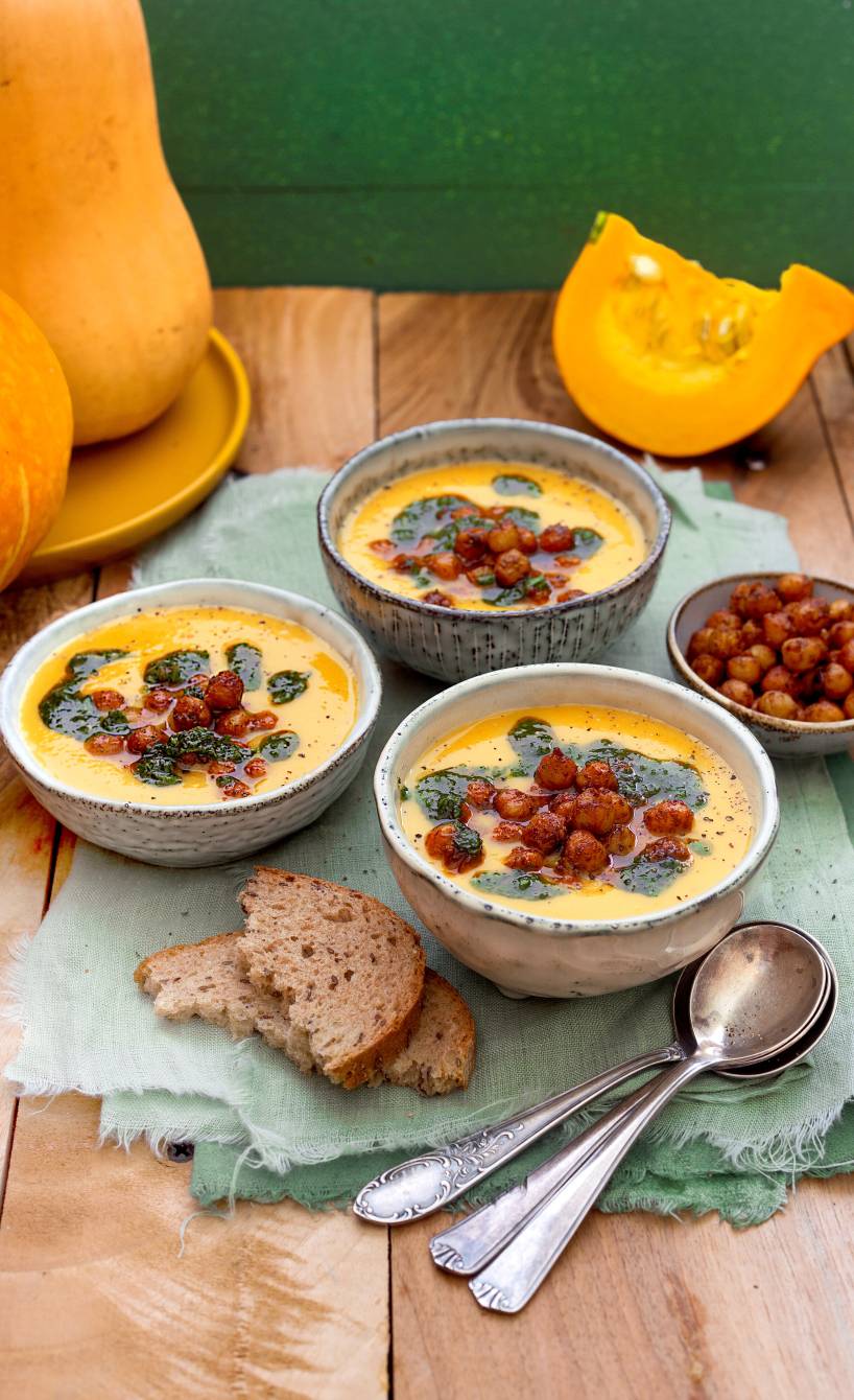 Selbstgekochte Muskatkürbis-Suppe in drei Supperschalen getoppt mit Kichererbsen und Kräuteröl auf einem Holztisch