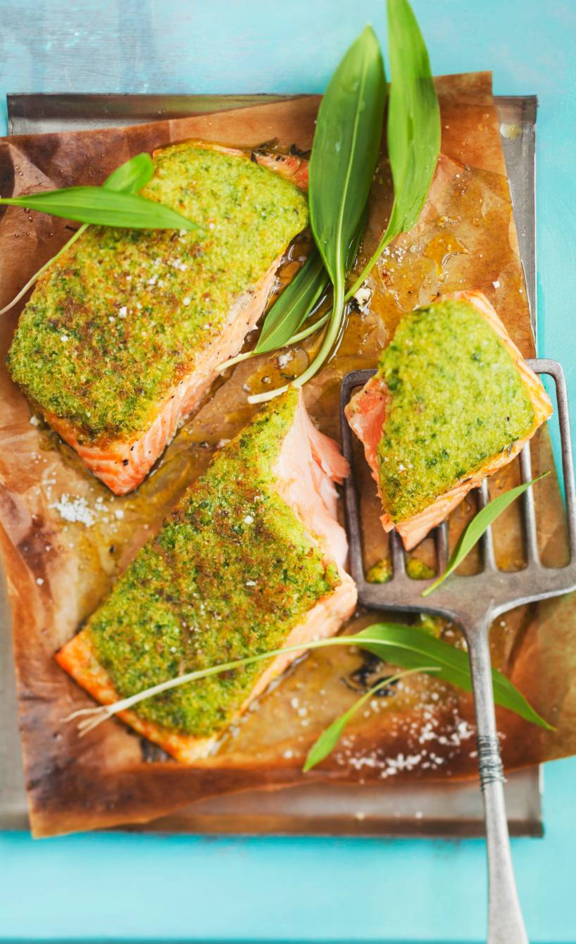 Mehrere Stücke gebackenes Lachsfilet mit Bärlauch-Parmesan-Kruste auf einem Backblech dekoriert mit frischen Bärlauchblättern