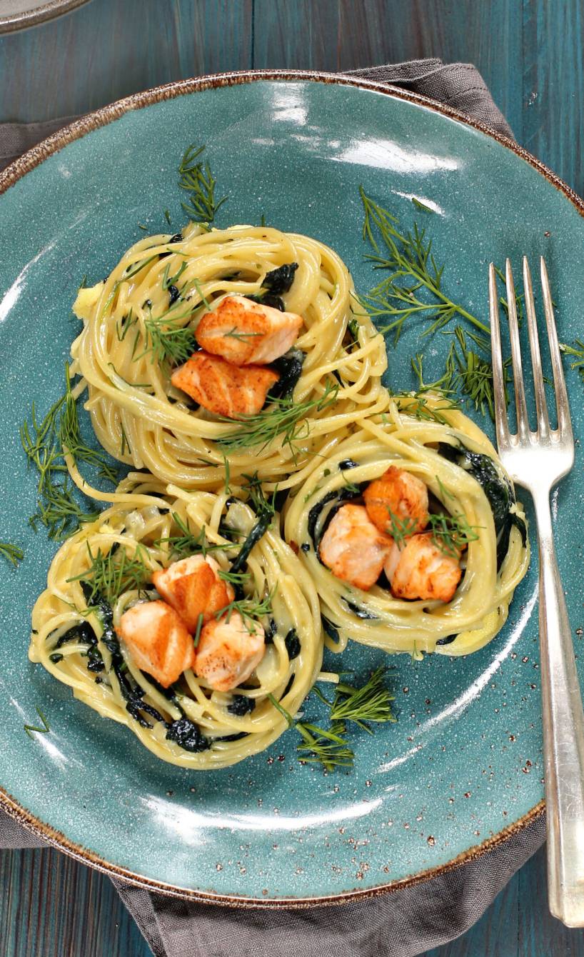 Drei Spaghettinester auf einem blauen Teller getoppt von Lachswürfeln und Spinat