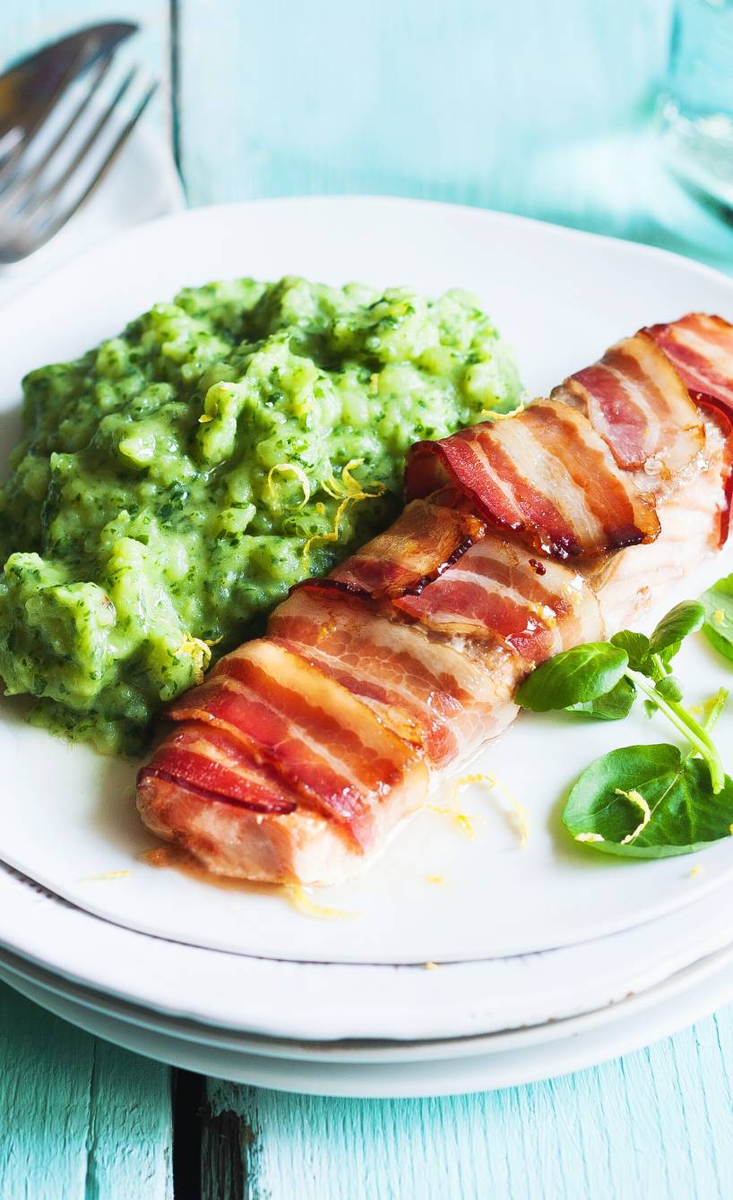Ein Stück Lachsfilet im Speckmantel auf einem Teller, daneben grüner Kartoffelstampf und frische Kresse